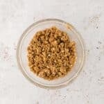 A glass bowl filled with brown crumbly mixture, possibly streusel or crumble topping, on a light-colored surface.
