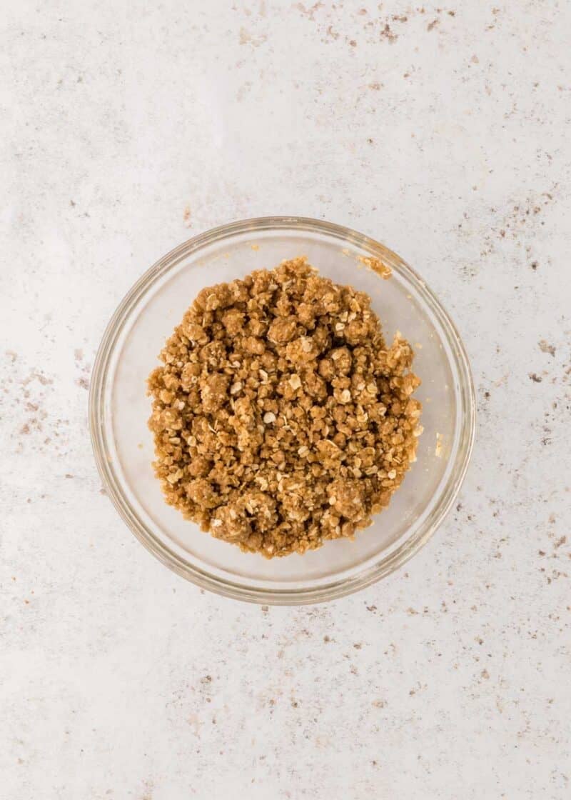 A glass bowl filled with brown crumbly mixture, possibly streusel or crumble topping, on a light-colored surface.