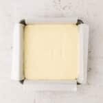 A square pan lined with parchment paper is filled with a smooth, pale batter, viewed from above on a light, textured surface.