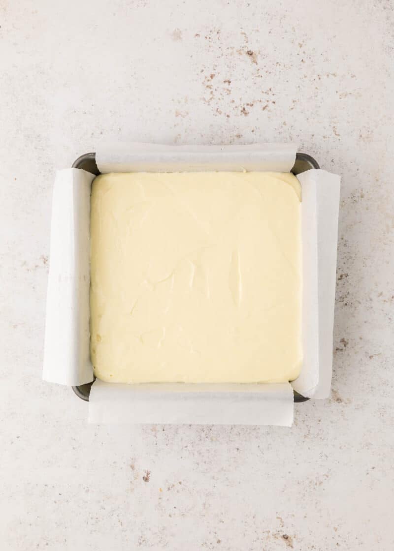 A square pan lined with parchment paper is filled with a smooth, pale batter, viewed from above on a light, textured surface.
