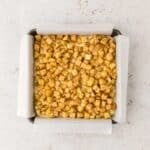 A square baking pan lined with parchment paper, filled with evenly spread, diced, seasoned apples on a light surface.