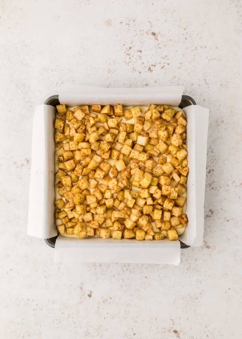 A square baking pan lined with parchment paper, filled with evenly spread, diced, seasoned apples on a light surface.