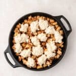 A black skillet filled with diced apples and topped with dollops of biscuit dough, ready to be baked.