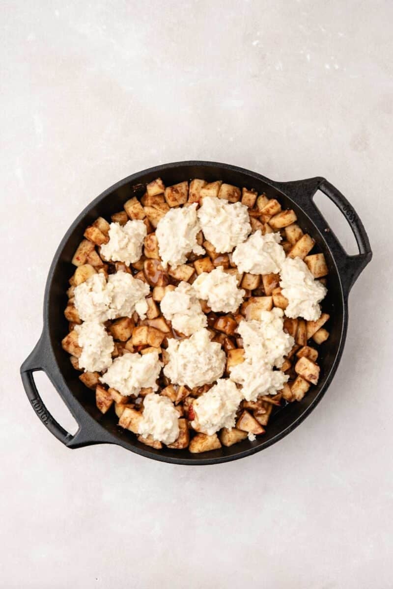 A black skillet filled with diced apples and topped with dollops of biscuit dough, ready to be baked.