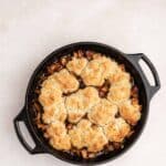 A black cast iron skillet filled with baked cobbler topped with golden-brown biscuit crust, sitting on a light-colored surface.