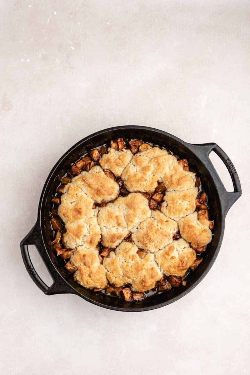 A black cast iron skillet filled with baked cobbler topped with golden-brown biscuit crust, sitting on a light-colored surface.