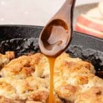 A wooden spoon drizzles caramel sauce over a golden-brown cobbler in a black skillet, with apple slices blurred in the background.