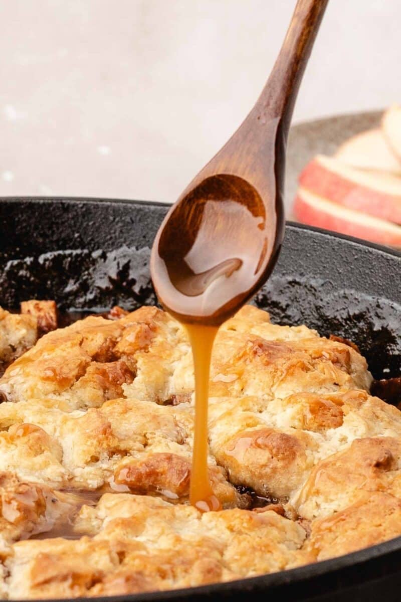 A wooden spoon drizzles caramel sauce over a golden-brown cobbler in a black skillet, with apple slices blurred in the background.