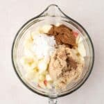 Glass bowl containing chopped apples, brown sugar, flour, and spices on a light surface, viewed from above.