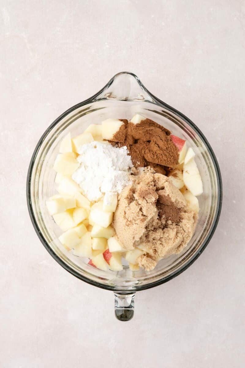 Glass bowl containing chopped apples, brown sugar, flour, and spices on a light surface, viewed from above.