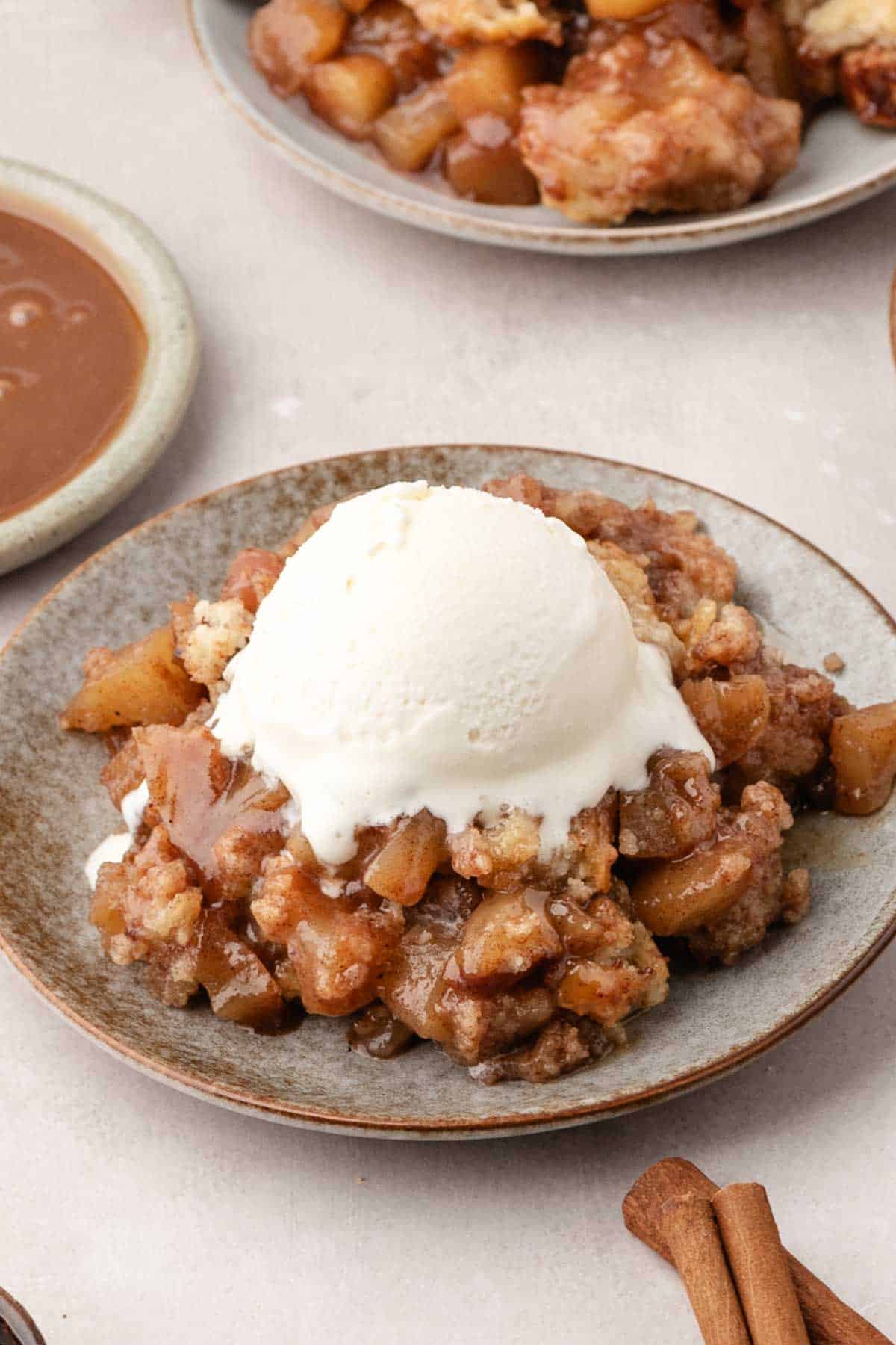 A plate of apple cobbler topped with a scoop of vanilla ice cream, with caramel sauce and cinnamon sticks nearby.