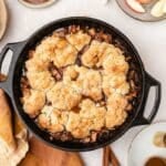 A cast iron skillet filled with baked apple cobbler, surrounded by plates with apple slices and spoons, on a light surface with a brown and beige napkin.