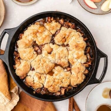 A cast iron skillet filled with baked apple cobbler, surrounded by plates with apple slices and spoons, on a light surface with a brown and beige napkin.