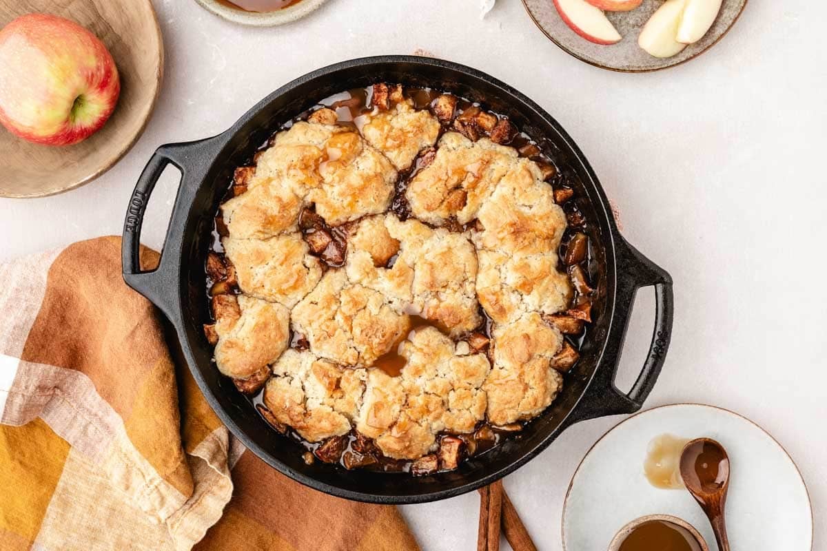 A cast iron skillet filled with baked apple cobbler, surrounded by plates with apple slices and spoons, on a light surface with a brown and beige napkin.
