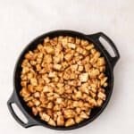 Cubed, seasoned potatoes are spread evenly in a black cast iron skillet on a light-colored surface.