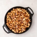 A black cast iron skillet filled with cubed apples coated in cinnamon and a brown sauce, placed on a light-colored surface.