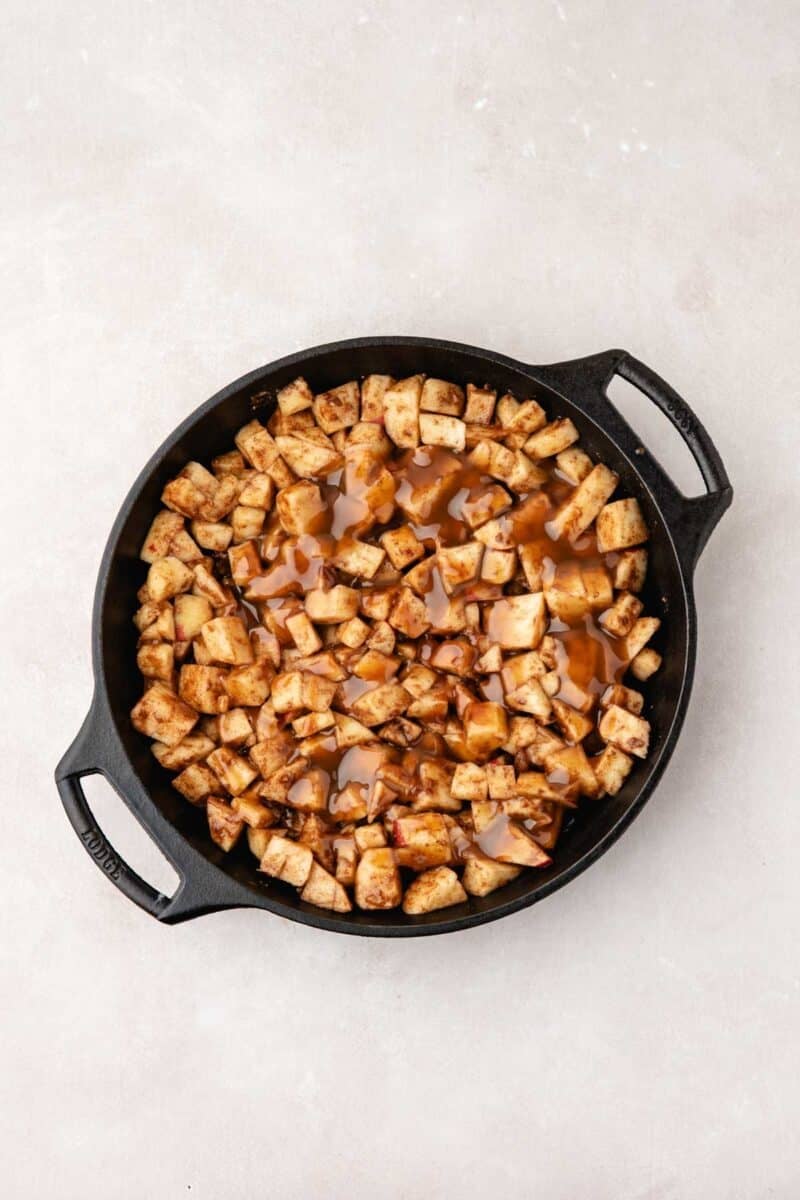 A black cast iron skillet filled with cubed apples coated in cinnamon and a brown sauce, placed on a light-colored surface.