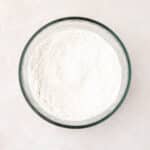 A glass bowl filled with white flour sits on a light-colored surface.