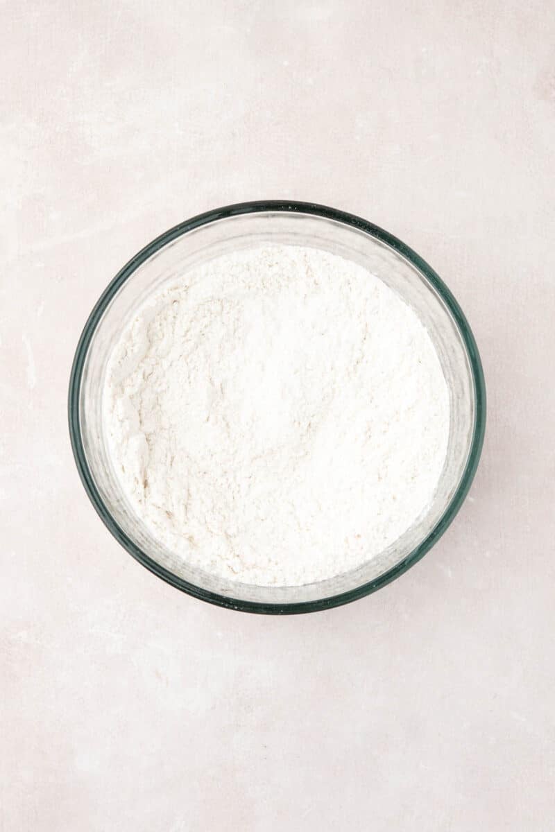 A glass bowl filled with white flour sits on a light-colored surface.