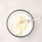 A glass bowl contains flour and several pieces of butter, with a metal fork resting inside, on a light-colored surface.