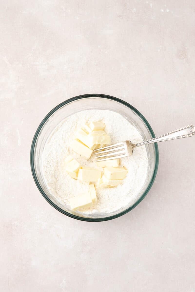 A glass bowl contains flour and several pieces of butter, with a metal fork resting inside, on a light-colored surface.