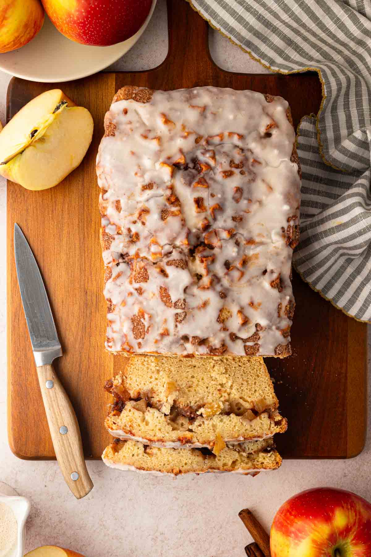 A loaf of glazed apple bread on a wooden cutting board, partially sliced, with a knife, apples, and a striped cloth nearby.
