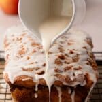 A hand pours white glaze from a small pitcher onto a loaf cake resting on a wire rack, with apples blurred in the background.