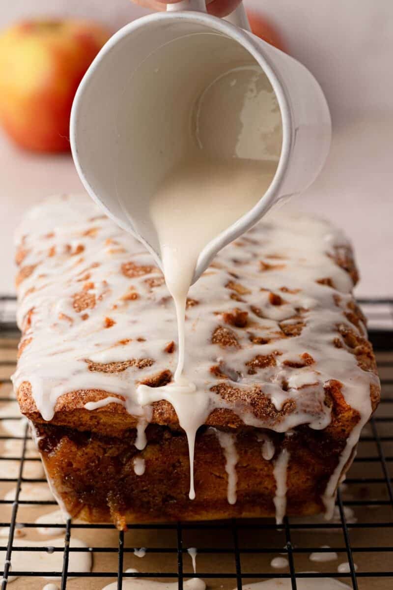 A hand pours white glaze from a small pitcher onto a loaf cake resting on a wire rack, with apples blurred in the background.