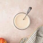 Glass bowl of creamy sauce with a spoon, next to two apples and a gray-striped cloth on a light countertop.