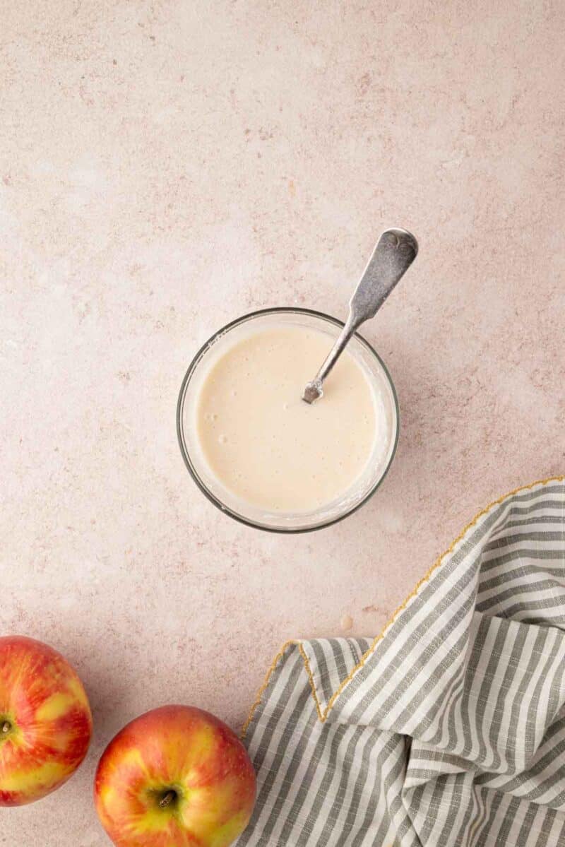 Glass bowl of creamy sauce with a spoon, next to two apples and a gray-striped cloth on a light countertop.