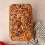 A loaf of apple bread with a cinnamon sugar topping sits on parchment paper next to two apples and a striped kitchen towel on a light countertop.