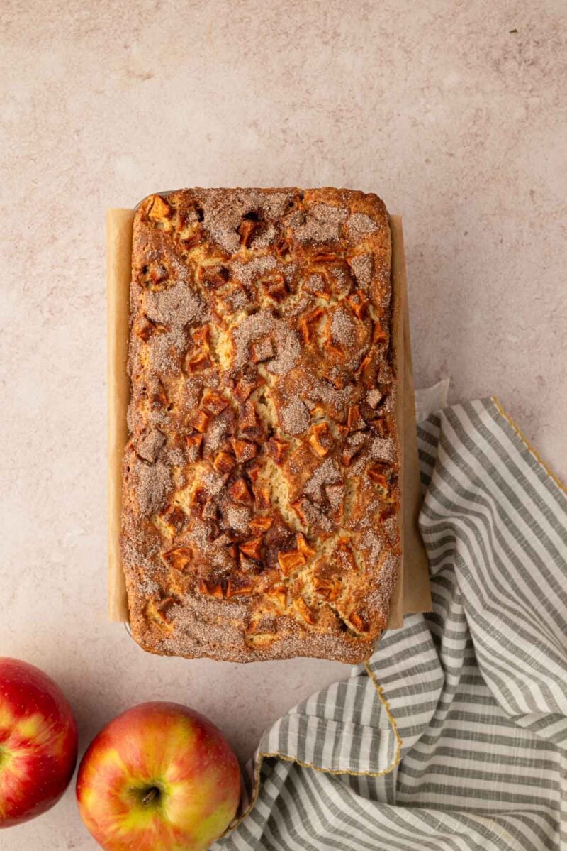 A loaf of apple bread with a cinnamon sugar topping sits on parchment paper next to two apples and a striped kitchen towel on a light countertop.