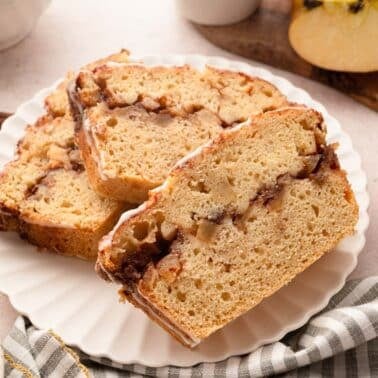 Three slices of apple cinnamon bread are arranged on a white plate, with apples, cinnamon sticks, and a striped cloth napkin nearby.