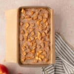 A rectangular baking dish lined with parchment paper holds a baked apple dessert topped with cinnamon and diced apples. Two apples and a striped cloth are beside the dish on a light surface.