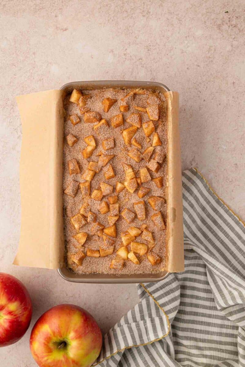 A rectangular baking dish lined with parchment paper holds a baked apple dessert topped with cinnamon and diced apples. Two apples and a striped cloth are beside the dish on a light surface.