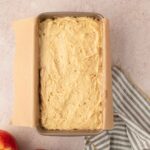 A loaf pan lined with parchment paper is filled with unbaked batter, with two apples and a striped cloth nearby on a light surface.