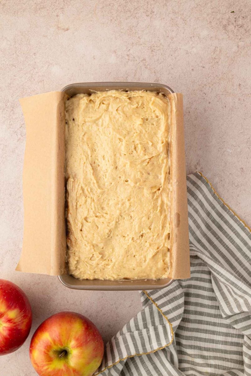 A loaf pan lined with parchment paper is filled with unbaked batter, with two apples and a striped cloth nearby on a light surface.