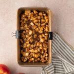 A parchment-lined loaf pan filled with diced apples mixed with cinnamon sits on a counter next to two apples and a striped cloth.
