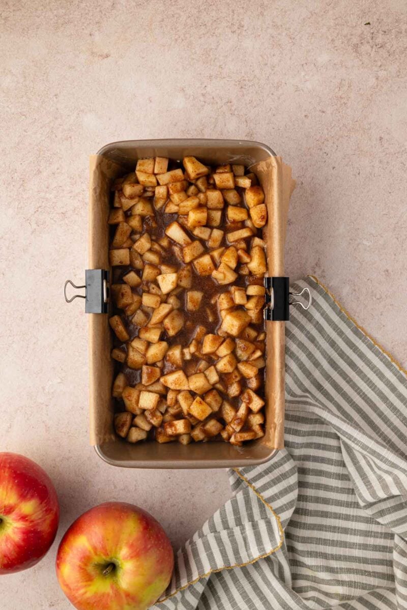 A parchment-lined loaf pan filled with diced apples mixed with cinnamon sits on a counter next to two apples and a striped cloth.
