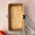 A loaf pan lined with parchment paper filled with unbaked batter, next to two apples and a striped kitchen towel on a light surface.