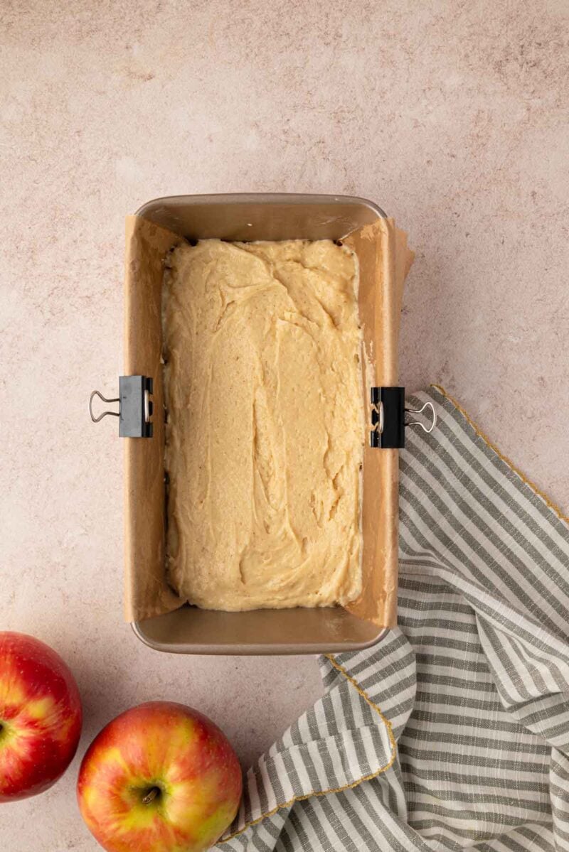 A loaf pan lined with parchment paper filled with unbaked batter, next to two apples and a striped kitchen towel on a light surface.