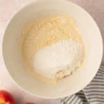 A white mixing bowl with cake batter and flour, next to two apples and a striped cloth on a light countertop.