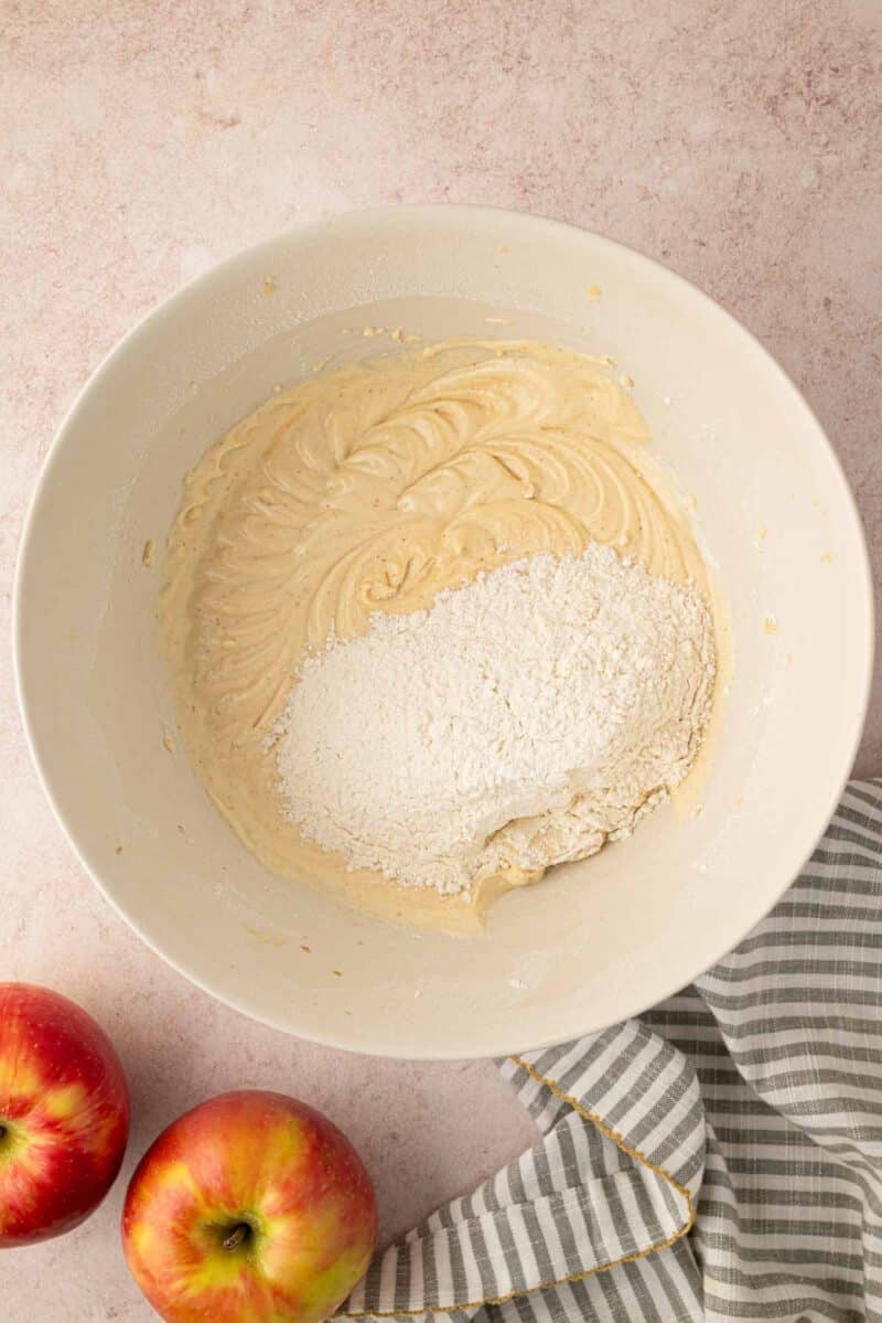 A white mixing bowl with cake batter and flour, next to two apples and a striped cloth on a light countertop.
