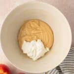 A mixing bowl with thick batter and a dollop of white yogurt, next to two apples and a striped kitchen towel on a light countertop.