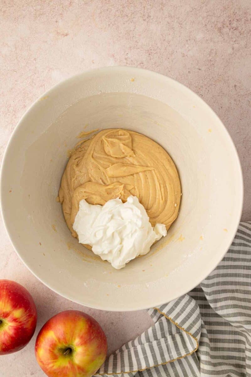 A mixing bowl with thick batter and a dollop of white yogurt, next to two apples and a striped kitchen towel on a light countertop.