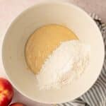 A mixing bowl contains batter and flour, sitting on a striped cloth with two apples nearby on a light countertop.