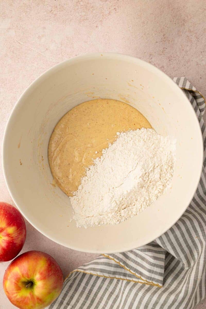 A mixing bowl contains batter and flour, sitting on a striped cloth with two apples nearby on a light countertop.
