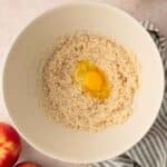 A mixing bowl with creamed ingredients and a cracked egg in the center, next to apples, a striped towel, and eggshells on a light surface.