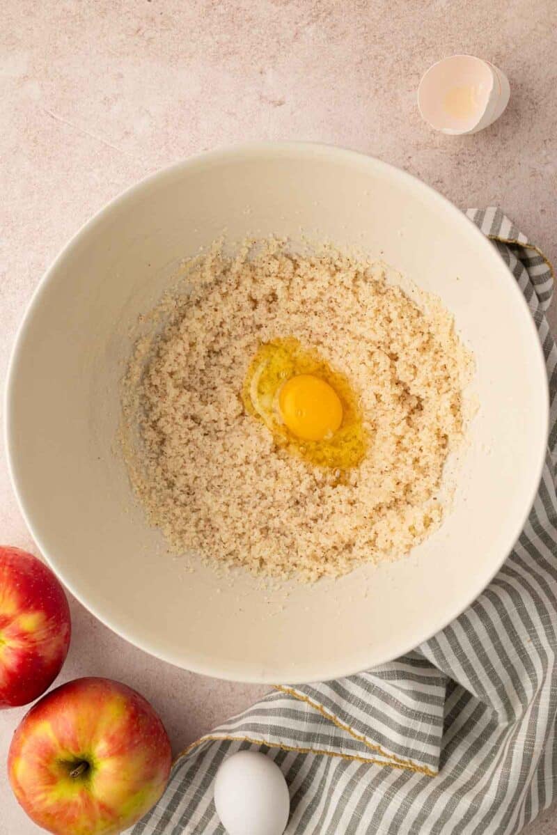 A mixing bowl with creamed ingredients and a cracked egg in the center, next to apples, a striped towel, and eggshells on a light surface.