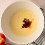 A mixing bowl with batter and a splash of vanilla extract, next to two apples, an empty small bowl, and a striped cloth on a light surface.
